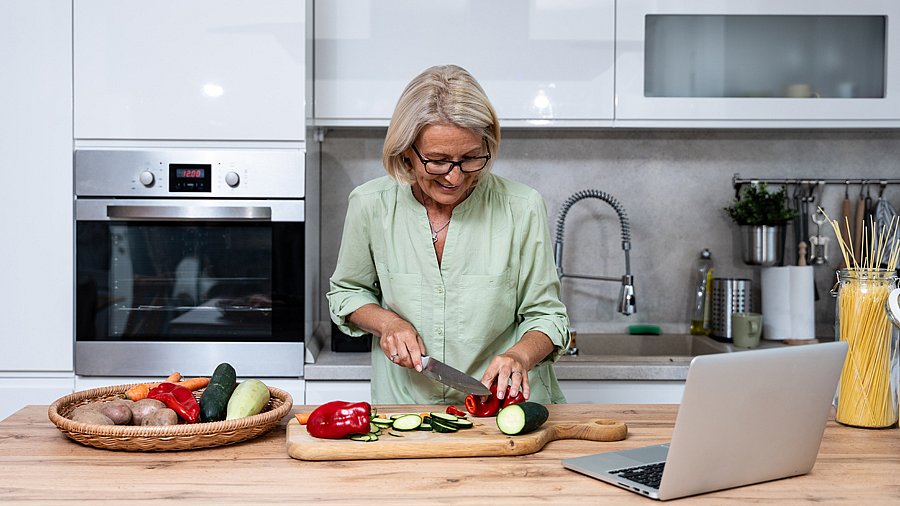 Frau steht in einer Küche und schneidet Gemüse, vor ihr ein aufgeklappter Laptop